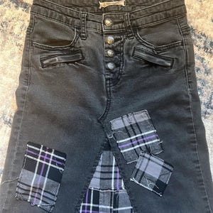 May include: Dark gray denim skirt with a button fly and decorative pockets. The skirt features three plaid patches in black, gray, and purple. The patches are sewn onto the skirt, adding a unique, distressed look.