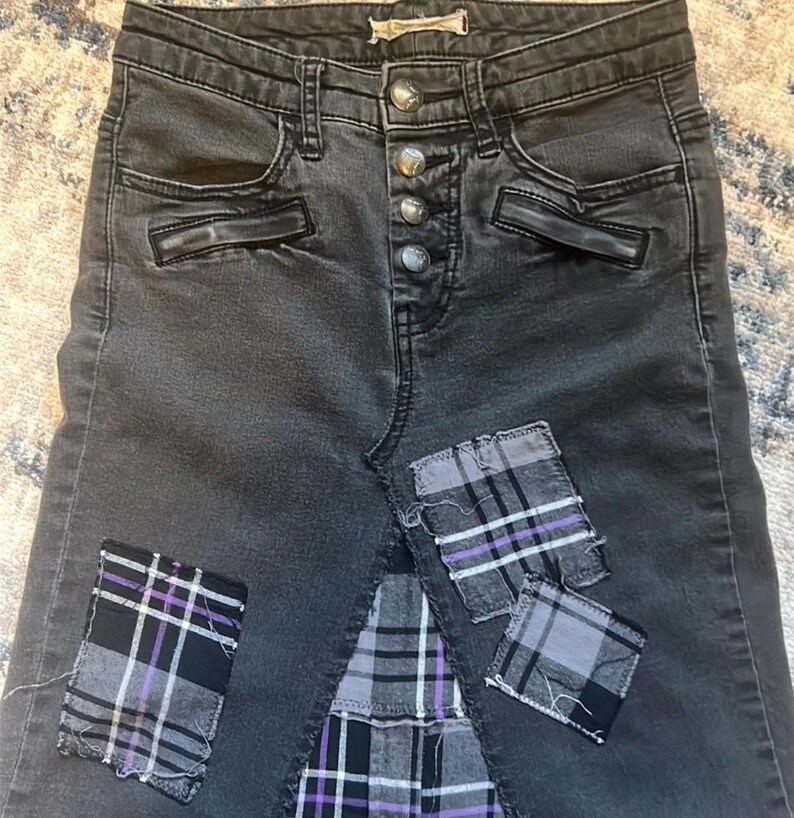 May include: Dark gray denim skirt with a button fly and decorative pockets. The skirt features three plaid patches in black, gray, and purple. The patches are sewn onto the skirt, adding a unique, distressed look.