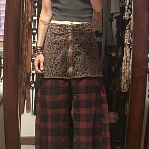 May include: A two-tone skirt with a leopard print top and a red and black plaid bottom. The skirt has a zipper closure and button details. The person in the image is wearing a dark gray t-shirt with a graphic design.