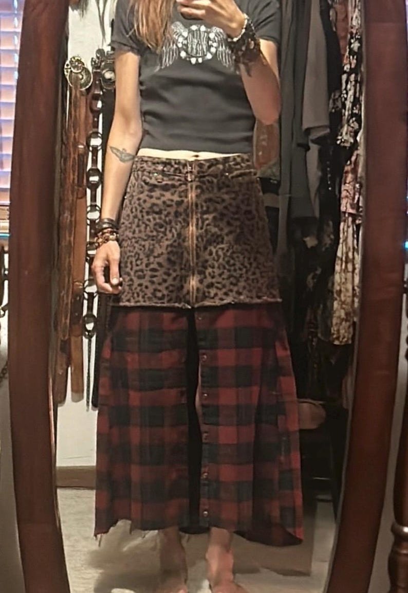 May include: A two-tone skirt with a leopard print top and a red and black plaid bottom. The skirt has a zipper closure and button details. The person in the image is wearing a dark gray t-shirt with a graphic design.