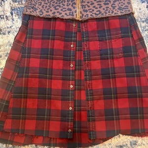May include: A red and black plaid skirt with a brown leopard print waistband. The skirt has a button-up front and a pocket on the right side. The skirt has a raw hemline.