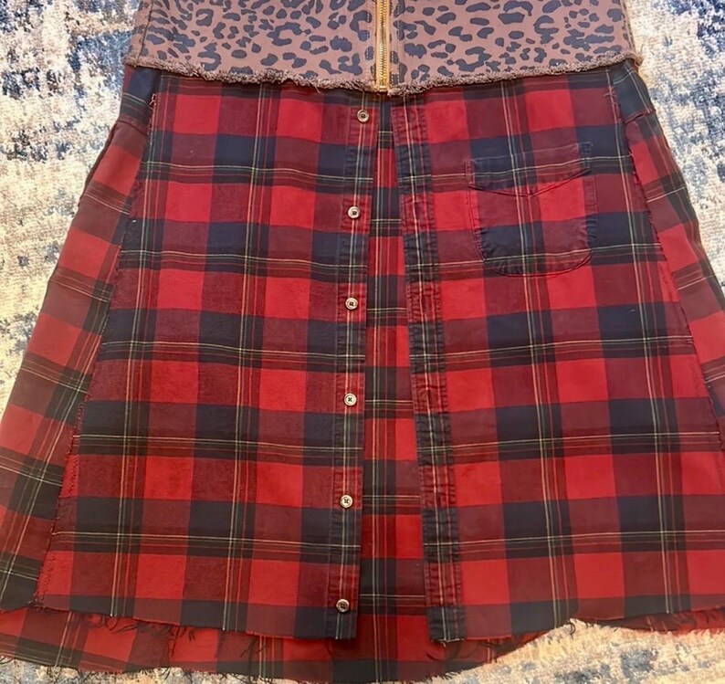 May include: A red and black plaid skirt with a brown leopard print waistband. The skirt has a button-up front and a pocket on the right side. The skirt has a raw hemline.