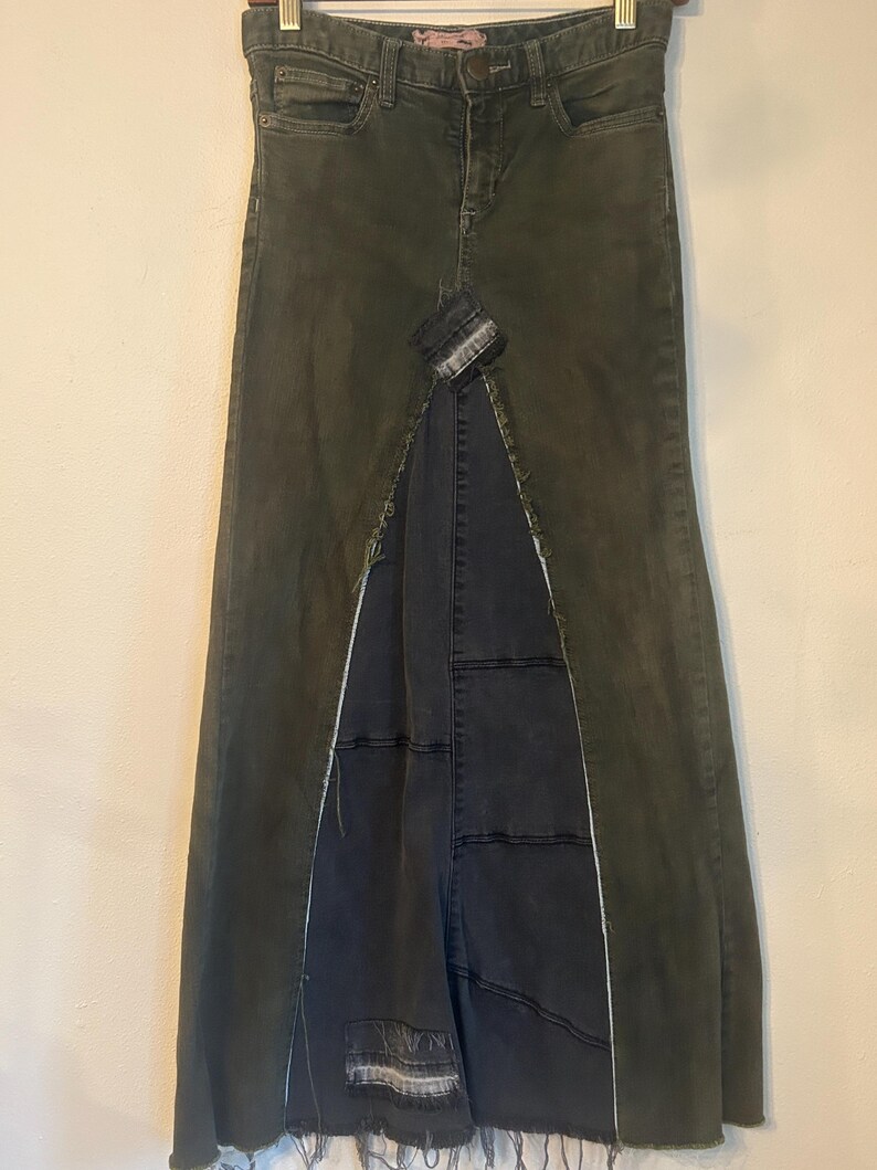 Puede incluir: Una falda larga de mezclilla verde oliva con una abertura frontal, que revela un panel de mezclilla gris m&aacute;s oscuro. La falda presenta un dise&ntilde;o de patchwork y un dobladillo deshilachado, que ofrece una est&eacute;tica &uacute;nica y desgastada. La falda tiene un dise&ntilde;o cl&aacute;sico de cinco bolsillos.