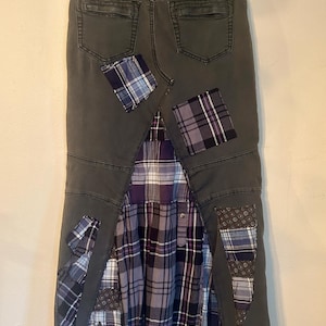 Puede incluir: Falda vaquera gris oscuro con detalles de patchwork. La falda presenta un inserto de tela a cuadros plisada en tonos morado, gris y blanco. Parches de tela a cuadros y estampada est&aacute;n cosidos en la mezclilla, agregando un aspecto &uacute;nico y artesanal.