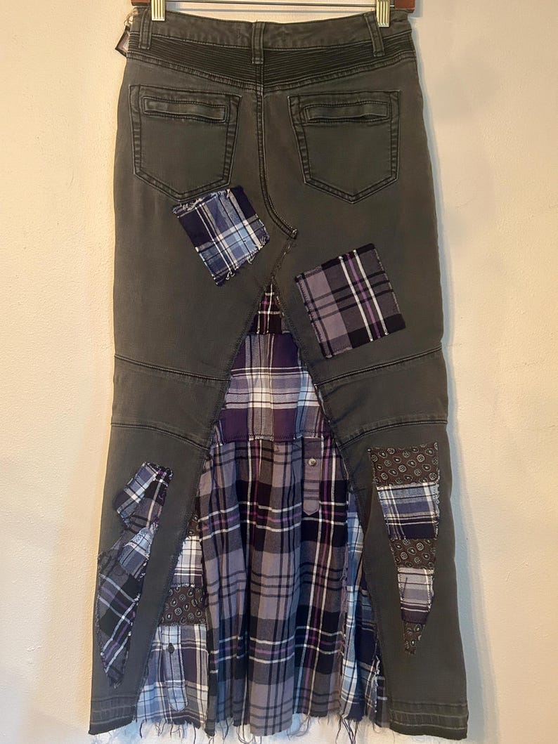 Puede incluir: Falda vaquera gris oscuro con detalles de patchwork. La falda presenta un inserto de tela a cuadros plisada en tonos morado, gris y blanco. Parches de tela a cuadros y estampada est&aacute;n cosidos en la mezclilla, agregando un aspecto &uacute;nico y artesanal.