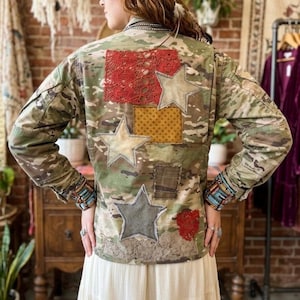 Könnte beinhalten: Eine Jacke mit Camouflage-Muster und verschiedenen Aufnähern auf der Rückseite, darunter drei Stoffsterne, ein rotes Häkelrechteck, ein senfgelbes Quadrat und ein dunkelbrauner Flicken. Die Ärmelbündchen sind mit bunten Perlen verziert.