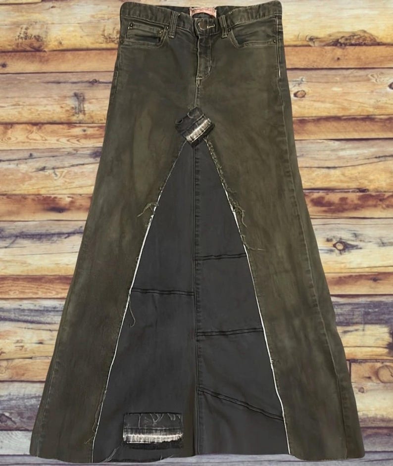 Puede incluir: Falda larga de mezclilla verde oliva con un dise&ntilde;o &uacute;nico. La falda presenta una abertura frontal con un panel gris m&aacute;s oscuro, costuras blancas y un aspecto desgastado. Tiene un dise&ntilde;o cl&aacute;sico de cinco bolsillos.