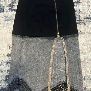 K&ouml;nnte beinhalten: Schwarzer Jeansrock mit silbernem Rei&szlig;verschluss und grauem Spitzeneinsatz. Der Rock hat ein einzigartiges Design mit geteilter Front und dekorativen Kn&ouml;pfen. Der untere Saum hat eine schwarze Spitzenborte.