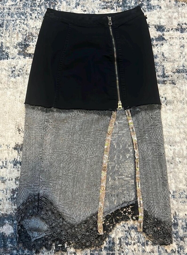 K&ouml;nnte beinhalten: Schwarzer Jeansrock mit silbernem Rei&szlig;verschluss und grauem Spitzeneinsatz. Der Rock hat ein einzigartiges Design mit geteilter Front und dekorativen Kn&ouml;pfen. Der untere Saum hat eine schwarze Spitzenborte.