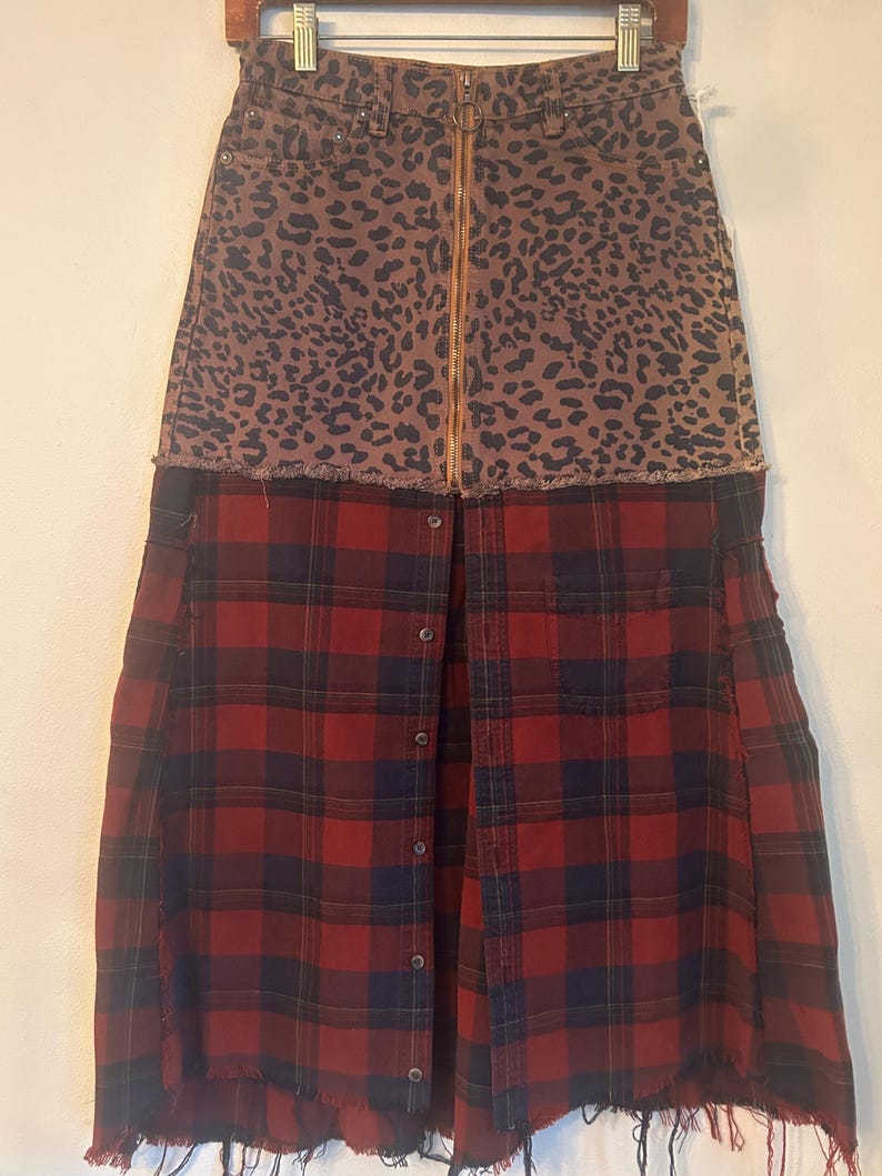 May include: A unique skirt featuring a leopard print denim top with a gold zipper and a red and black plaid flannel bottom. The skirt has a raw hem and button details.