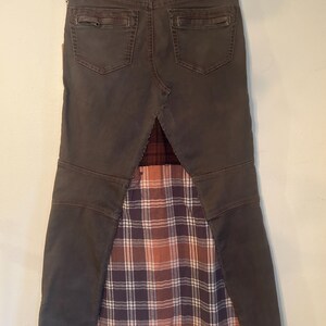 May include: A dark gray denim skirt with a plaid fabric panel. The skirt has two back pockets and a decorative stitched waistband. The plaid fabric features a mix of brown, orange, and purple hues.