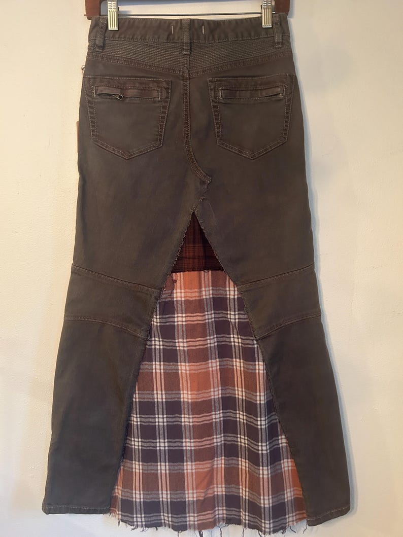 May include: A dark gray denim skirt with a plaid fabric panel. The skirt has two back pockets and a decorative stitched waistband. The plaid fabric features a mix of brown, orange, and purple hues.