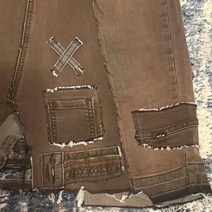 May include: Close-up of brown denim shorts with distressed details. Features include multiple pockets, decorative patches, and a raw hem. The shorts have an X-shaped patch and a patchwork design, giving them a unique, vintage aesthetic.