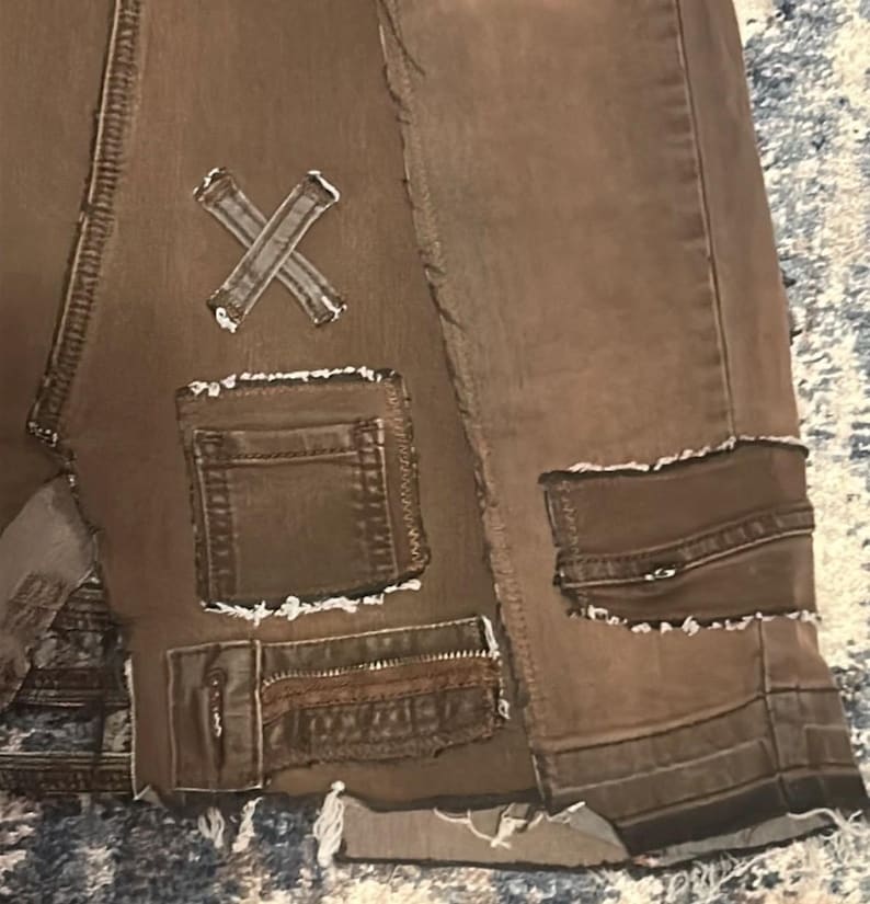 May include: Close-up of brown denim shorts with distressed details. Features include multiple pockets, decorative patches, and a raw hem. The shorts have an X-shaped patch and a patchwork design, giving them a unique, vintage aesthetic.