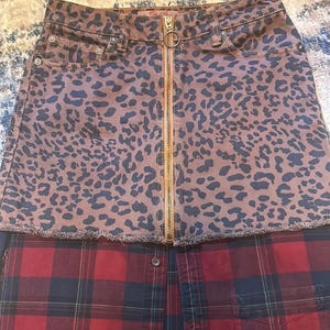 May include: A two-tone skirt featuring a brown leopard print denim top with a gold zipper and a red and black plaid bottom. The skirt has a raw hemline and a button closure.