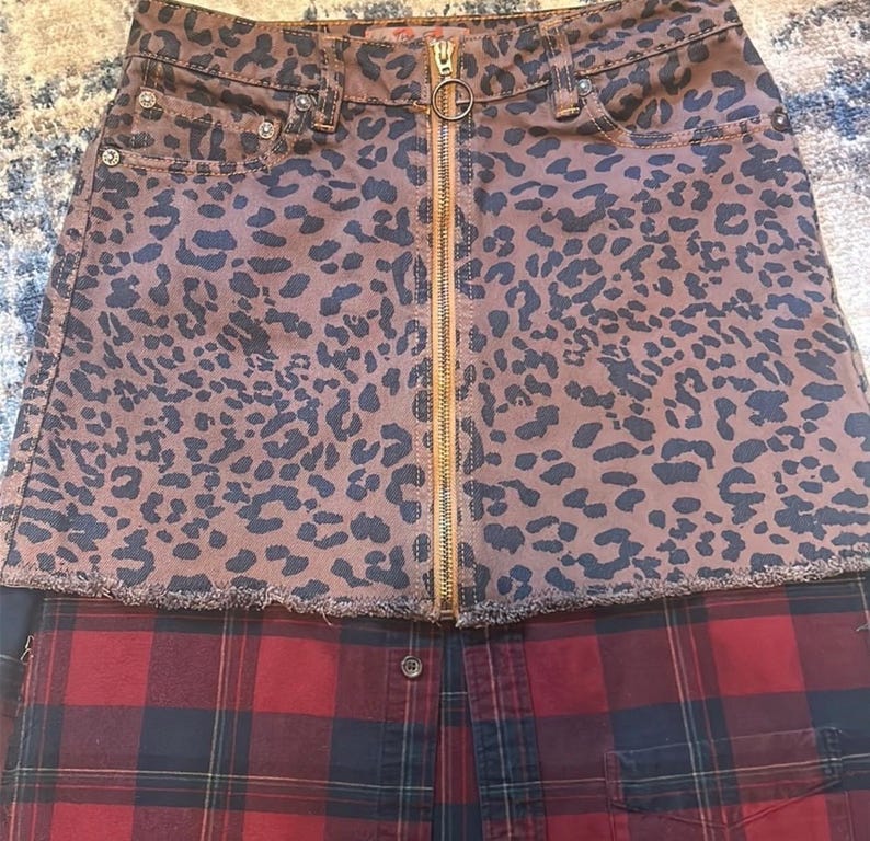 May include: A two-tone skirt featuring a brown leopard print denim top with a gold zipper and a red and black plaid bottom. The skirt has a raw hemline and a button closure.