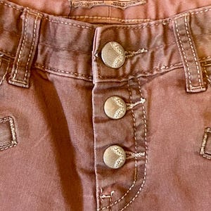 May include: Close-up of dusty rose-colored pants with three decorative buttons. The pants have white stitching and a label that reads "FREE PEOPLE" and "SIZE: W 25".