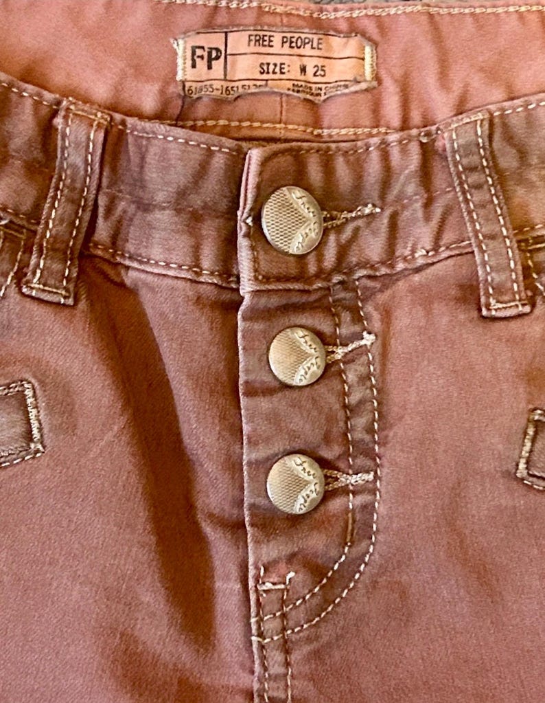 May include: Close-up of dusty rose-colored pants with three decorative buttons. The pants have white stitching and a label that reads "FREE PEOPLE" and "SIZE: W 25".