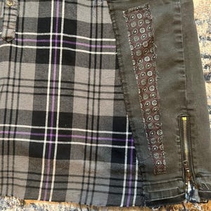 Puede incluir: Una falda a cuadros gris, negro y morado con un panel de mezclilla gris oscuro. El panel de mezclilla tiene un parche decorativo de cachemira y una cremallera dorada. La falda tiene un dobladillo sin rematar.