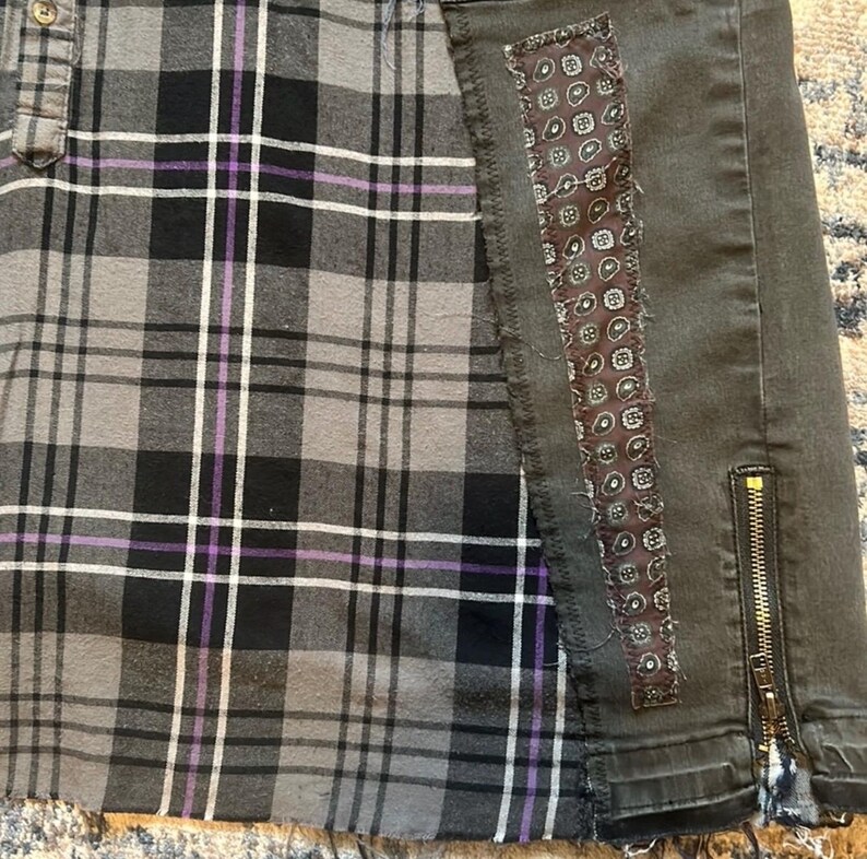 Puede incluir: Una falda a cuadros gris, negro y morado con un panel de mezclilla gris oscuro. El panel de mezclilla tiene un parche decorativo de cachemira y una cremallera dorada. La falda tiene un dobladillo sin rematar.