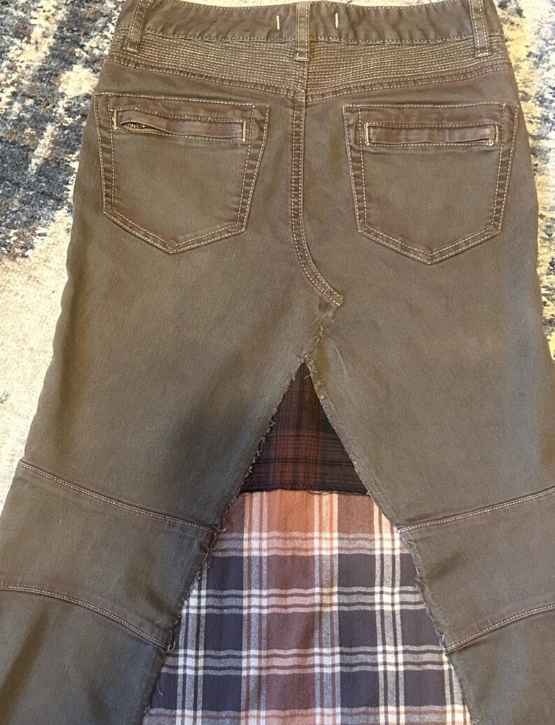 May include: A pair of olive green pants with two back pockets and a plaid fabric panel. The pants have a unique design with visible stitching and a contrasting plaid pattern at the bottom.