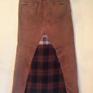May include: A brown denim skirt with a plaid fabric panel. The skirt features a distressed look with visible stitching and a frayed hem. The back pockets and waistband are visible.