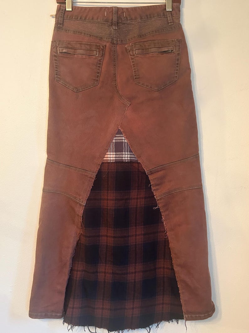 May include: A brown denim skirt with a plaid fabric panel. The skirt features a distressed look with visible stitching and a frayed hem. The back pockets and waistband are visible.