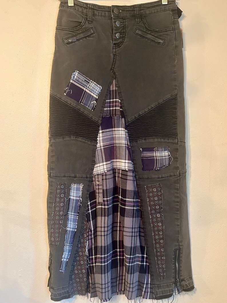 Puede incluir: Falda vaquera gris oscuro con detalles de patchwork. La falda presenta una mezcla de texturas, incluyendo paneles acolchados e inserciones de tela a cuadros en tonos morados, azules y grises. Tiene cierre de bot&oacute;n y cremallera, y costuras decorativas.