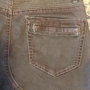 May include: Close-up of the back of brown pants with a pocket. The pants have white stitching along the seams and around the pocket. The waistband features horizontal stitching. The pocket is rectangular with a small, stitched detail.