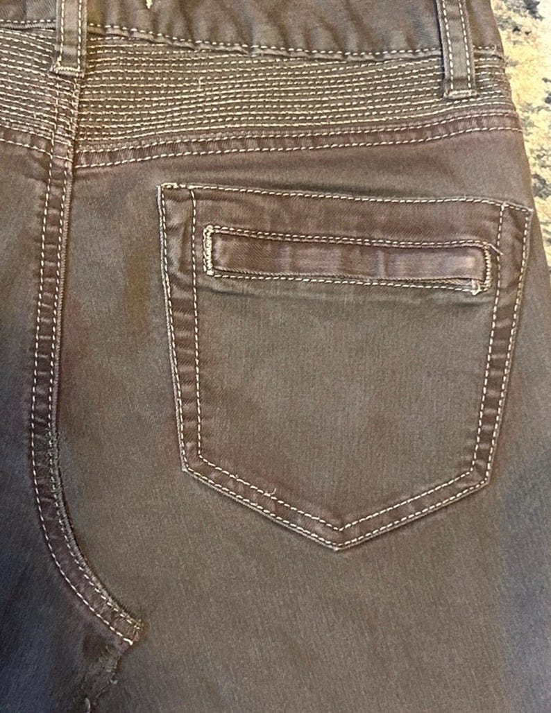 May include: Close-up of the back of brown pants with a pocket. The pants have white stitching along the seams and around the pocket. The waistband features horizontal stitching. The pocket is rectangular with a small, stitched detail.