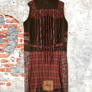 Może przedstawiać: Długa kamizelka bez rękawów z patchworkowym wzorem. Kamizelka ma ciemnobrązową górną część z czerwoną kratą i frędzlami z tyłu. Dolna część to czerwona krata z brązowymi łatami.