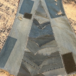May include: A denim skirt with a patchwork design. The skirt features various shades of blue denim, with patches and distressed details. Two black patches with silver studs are placed on the skirt. The skirt has a unique, asymmetrical shape.
