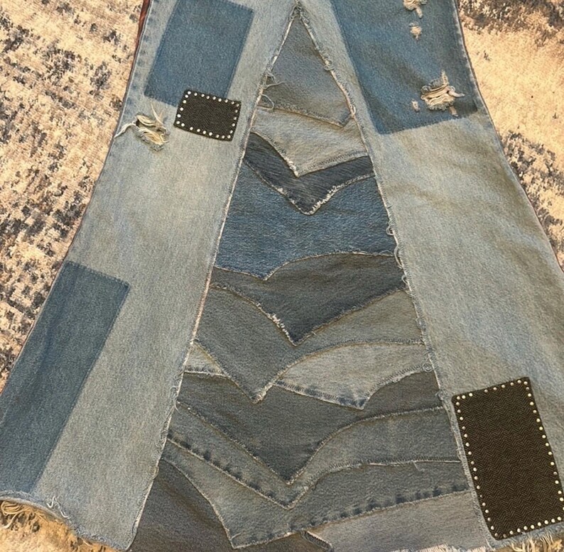 May include: A denim skirt with a patchwork design. The skirt features various shades of blue denim, with patches and distressed details. Two black patches with silver studs are placed on the skirt. The skirt has a unique, asymmetrical shape.