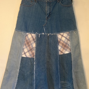 May include: A patchwork denim skirt with a light blue wash. The skirt features panels of different denim shades and plaid fabric. The skirt is hanging on a wooden hanger.