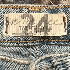 May include: Close-up of a light blue denim fabric with a rectangular tag. The tag reads "WE THE FREE 24 TWENTY FOUR REGULAR FABRIQU&Eacute; EN TURQUIE". The denim has visible stitching and a textured appearance.