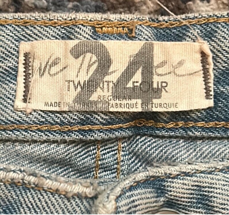 May include: Close-up of a light blue denim fabric with a rectangular tag. The tag reads "WE THE FREE 24 TWENTY FOUR REGULAR FABRIQU&Eacute; EN TURQUIE". The denim has visible stitching and a textured appearance.