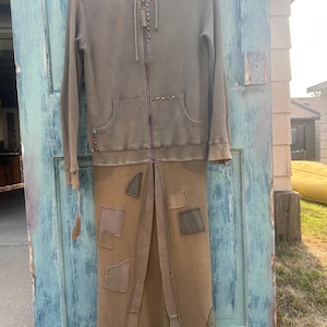 May include: A long-sleeved, hooded zip-up jacket in a muted olive-brown waffle knit fabric. The garment features a front zipper, a hood with drawstrings, and a long, skirt-like lower section adorned with various rectangular fabric patches in earth tones.