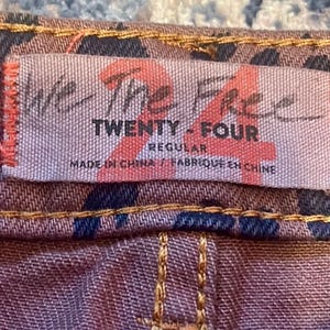 May include: Close-up of a tag on a pair of jeans. The tag reads "We The Free TWENTY - FOUR REGULAR MADE IN CHINA / FABRIQUE EN CHINE". The jeans have a brown and navy blue pattern with tan stitching.