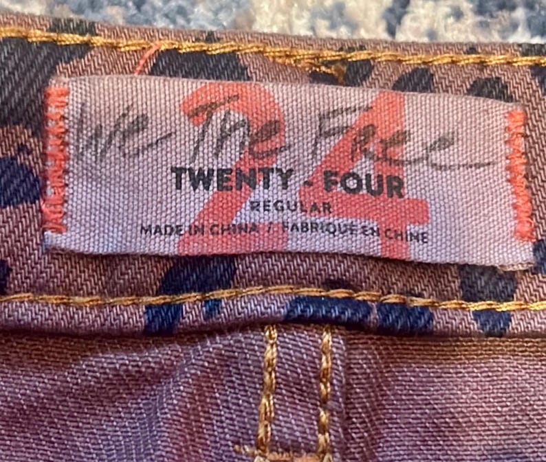 May include: Close-up of a tag on a pair of jeans. The tag reads "We The Free TWENTY - FOUR REGULAR MADE IN CHINA / FABRIQUE EN CHINE". The jeans have a brown and navy blue pattern with tan stitching.