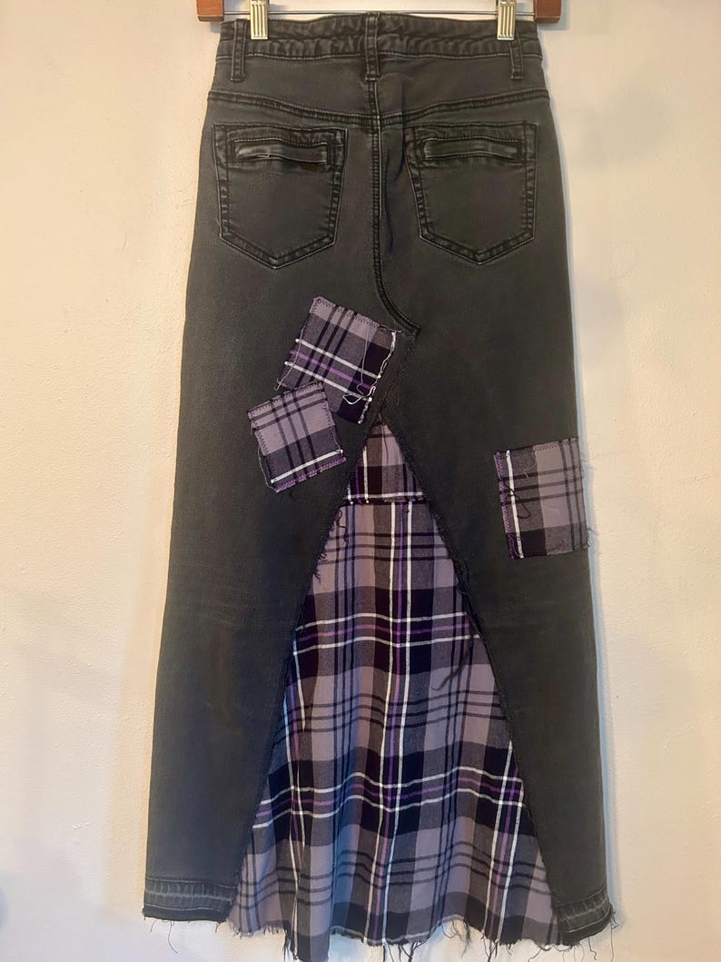May include: Dark gray denim skirt with a plaid fabric panel and patches. The skirt features a distressed hem and back pockets. The plaid fabric incorporates shades of purple, gray, and white, adding a unique design element to the garment.