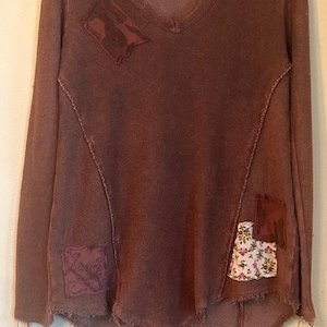 May include: A long-sleeve brown V-neck top featuring a high-low hemline with raw, frayed edges. The garment includes various fabric patches in solid brown, dark brown camouflage, and a white floral print. A tag labeled Sparrow 77 is attached.