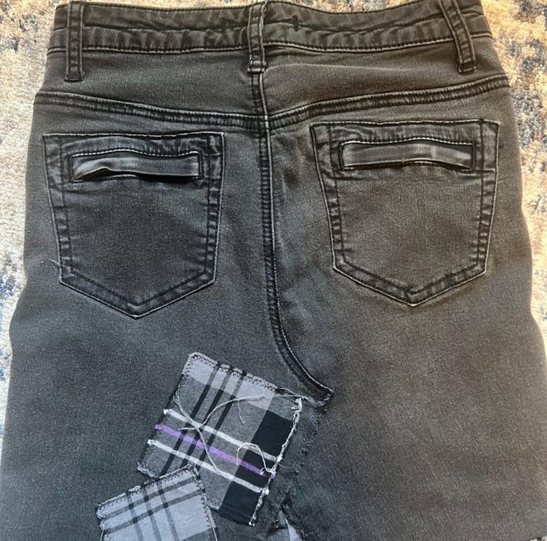 May include: Close-up of the back of a pair of dark gray denim shorts. The shorts have two back pockets and a small plaid patch sewn on the lower left side. The shorts are worn and faded.