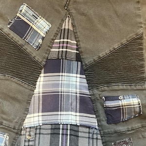 Puede incluir: Primer plano de unos pantalones vaqueros grises con detalles de patchwork. Los pantalones presentan un panel central de tela de cuadros en tonos azules, grises y morados. Parches de tela de cuadros est&aacute;n cosidos en la mezclilla, agregando un aspecto &uacute;nico y artesanal.