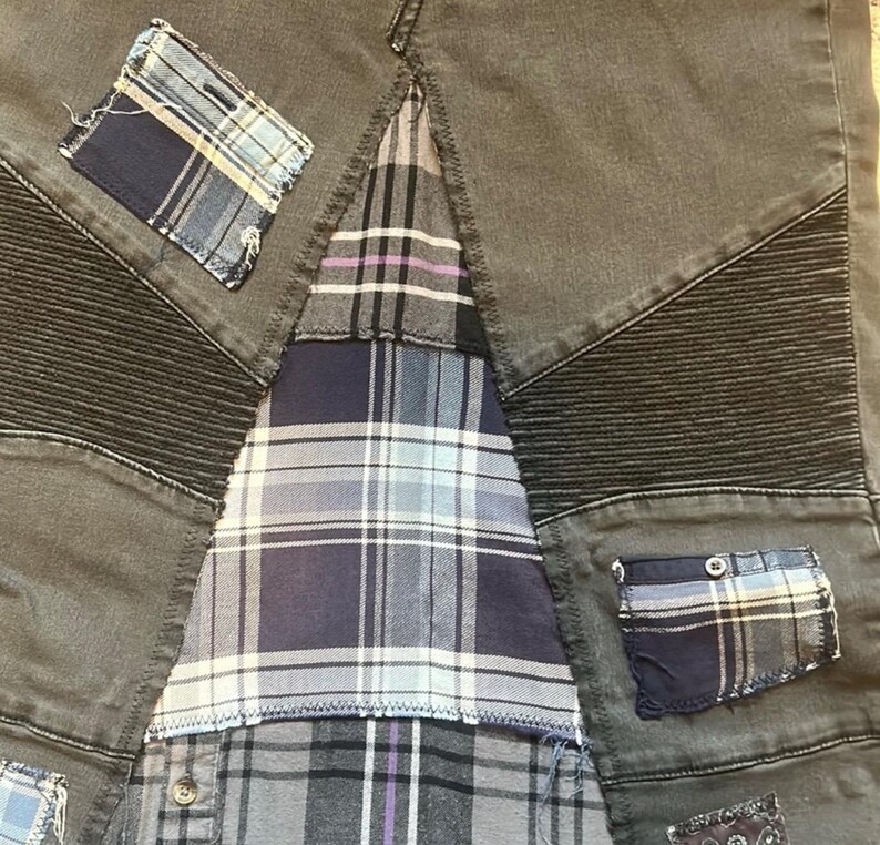 Puede incluir: Primer plano de unos pantalones vaqueros grises con detalles de patchwork. Los pantalones presentan un panel central de tela de cuadros en tonos azules, grises y morados. Parches de tela de cuadros est&aacute;n cosidos en la mezclilla, agregando un aspecto &uacute;nico y artesanal.
