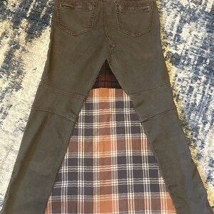 May include: A unique, repurposed skirt made from olive green denim and a plaid fabric insert. The skirt features the back pockets and waistband of the original jeans, with a flared, patchwork design. The plaid insert has brown, white, and navy blue tones.