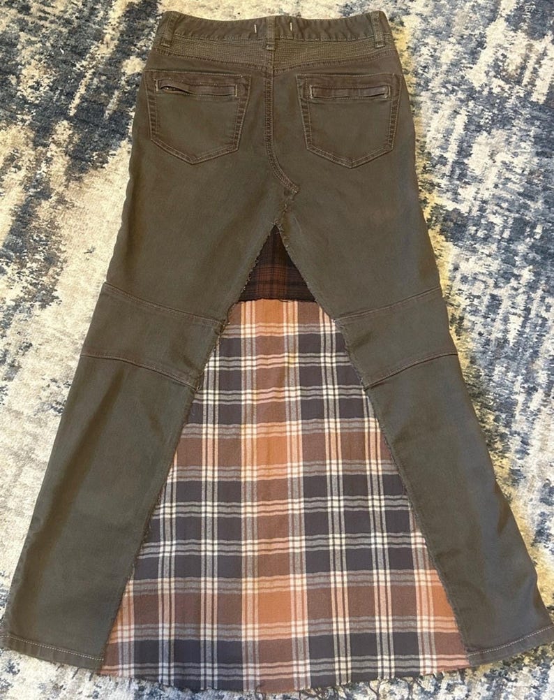 May include: A unique, repurposed skirt made from olive green denim and a plaid fabric insert. The skirt features the back pockets and waistband of the original jeans, with a flared, patchwork design. The plaid insert has brown, white, and navy blue tones.