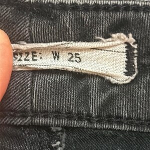 May include: Close-up of a black denim garment. A rectangular fabric tag displays the text "SIZE: W 25" in black lettering. The denim fabric has a textured appearance with visible stitching.