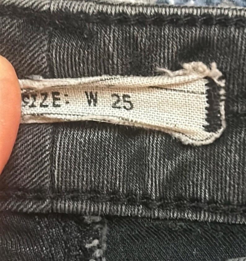 May include: Close-up of a black denim garment. A rectangular fabric tag displays the text "SIZE: W 25" in black lettering. The denim fabric has a textured appearance with visible stitching.