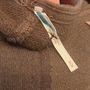 Może przedstawiać: Zbliżenie na brązowy sweter z dołączoną metką. Metka jest biała z niebieskimi i turkusowymi akcentami oraz napisem "MADE IN FABRIC". Sweter ma prążkowaną fakturę i wysoki dekolt. Na metce znajduje się r&oacute;wnież rozmiar "M".