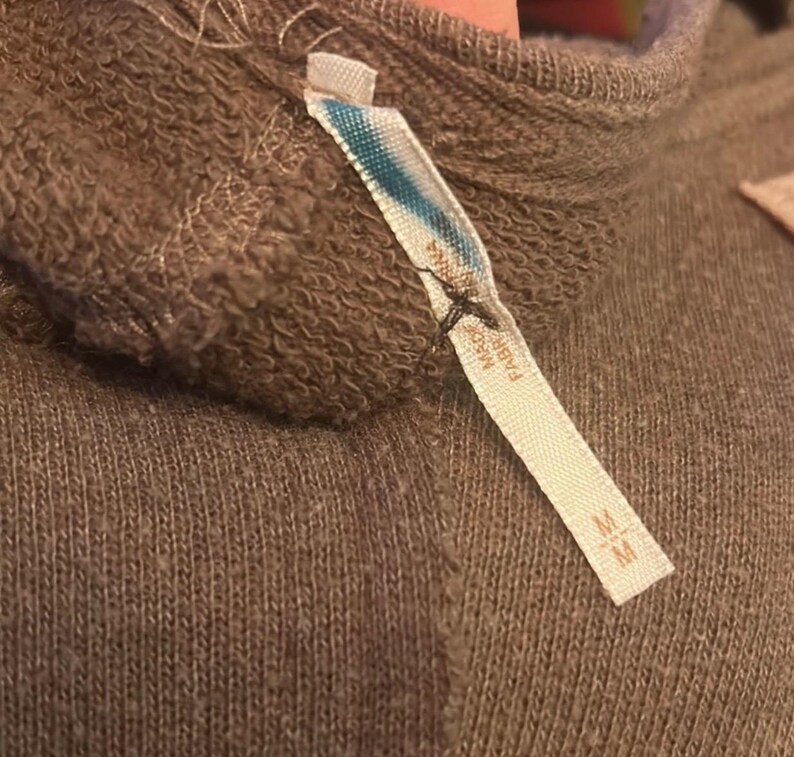 Może przedstawiać: Zbliżenie na brązowy sweter z dołączoną metką. Metka jest biała z niebieskimi i turkusowymi akcentami oraz napisem "MADE IN FABRIC". Sweter ma prążkowaną fakturę i wysoki dekolt. Na metce znajduje się r&oacute;wnież rozmiar "M".